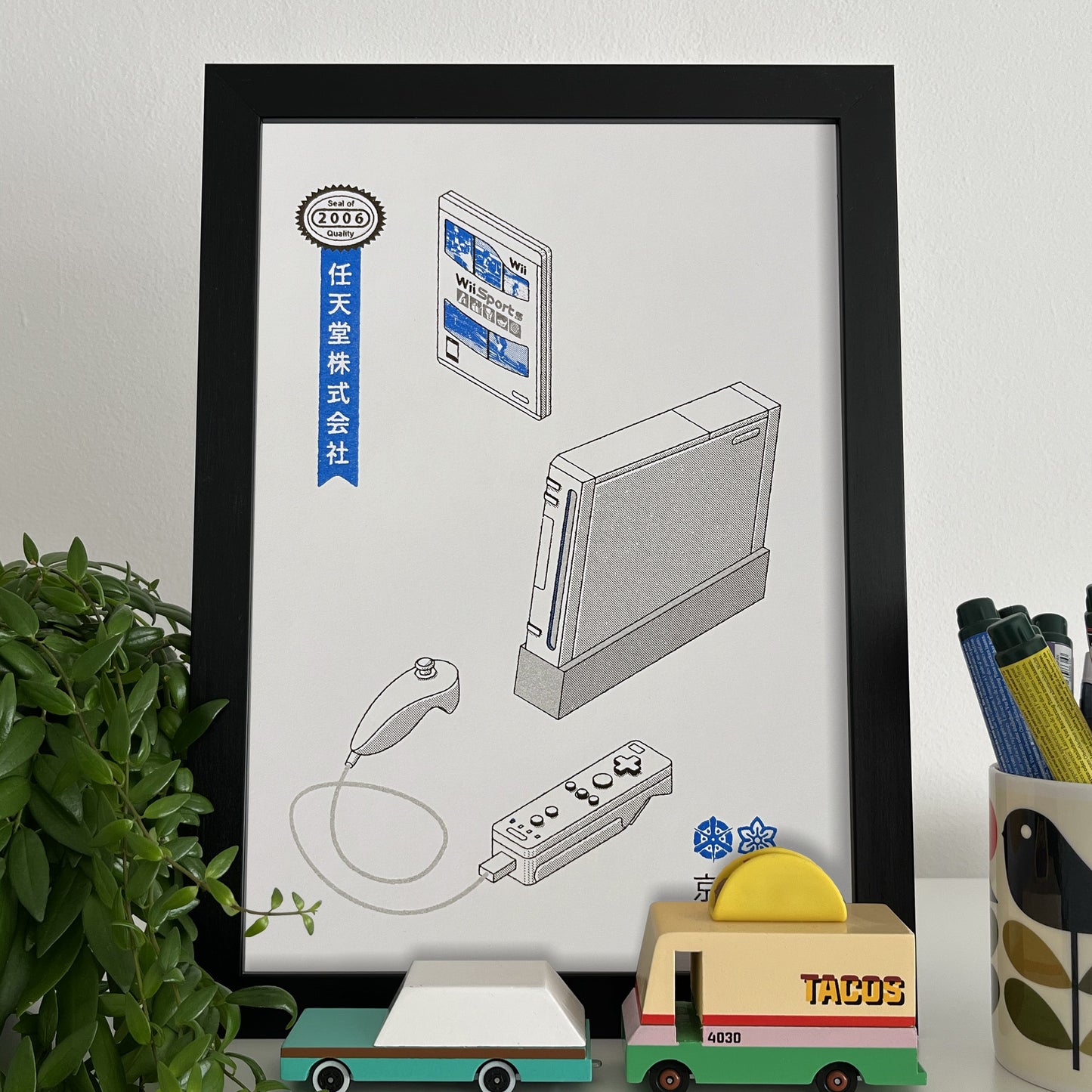 Framed illustration of a Nintendo Wii on a shelf with a plant and stationery items.